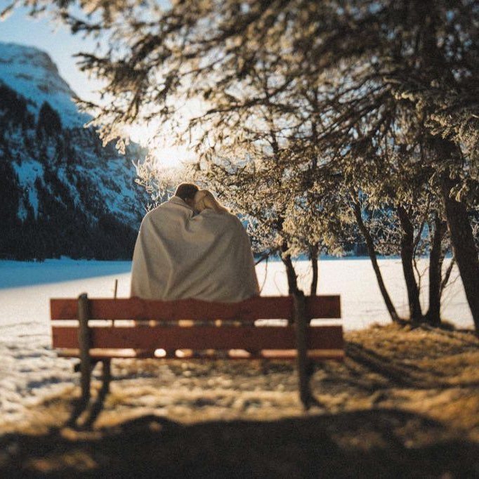 Paar in eine Decke gehüllt auf einer Bank am verschneiten Bergsee